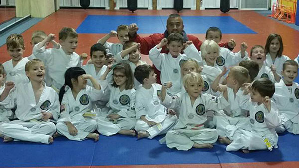 Taekwondo master kneels behind a large group of little ninja students with their fists in the air.
